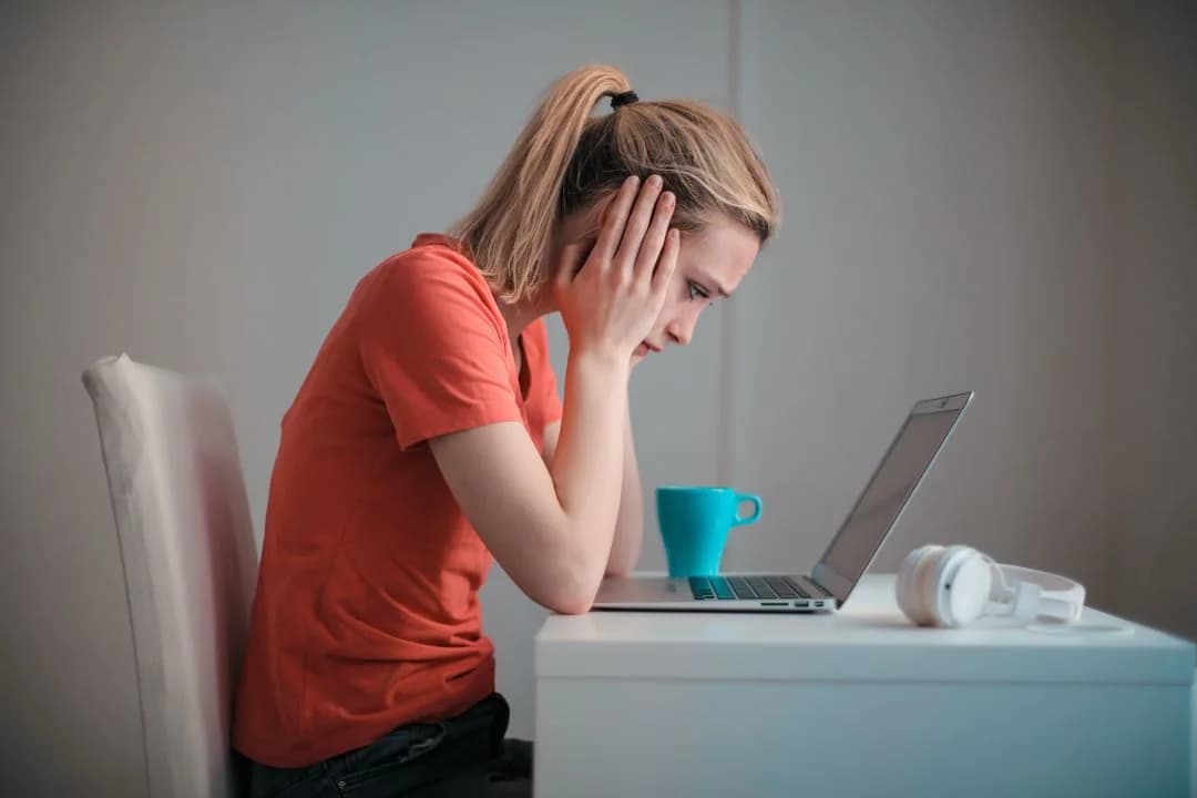 Stressed college student holding her head while studying at a laptop with a mug and headphones on the desk