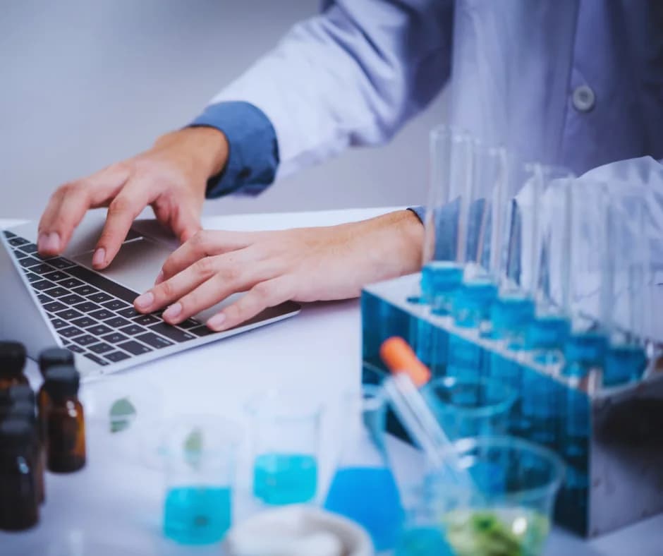 Scientist in a lab coat typing on a laptop beside racks of test tubes filled with blue liquid