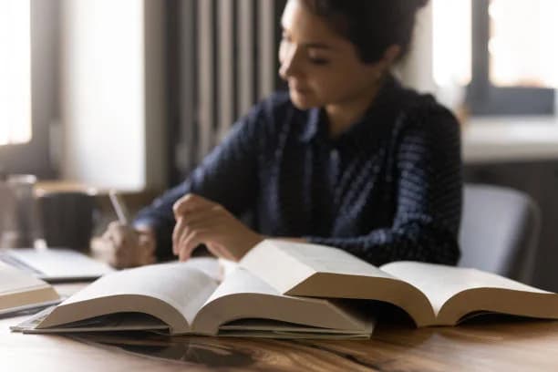 Graduate student taking notes at a wooden desk stacked with open books while working on her dissertation