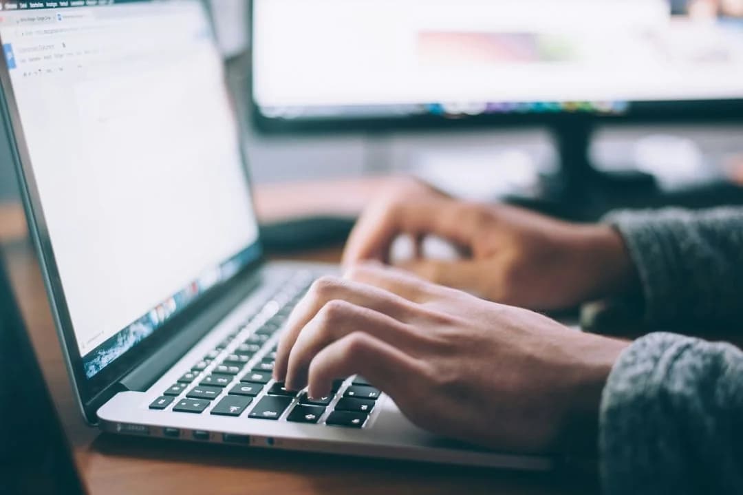 Close-up of hands typing on a laptop keyboard at a desk with a second monitor glowing in the background
