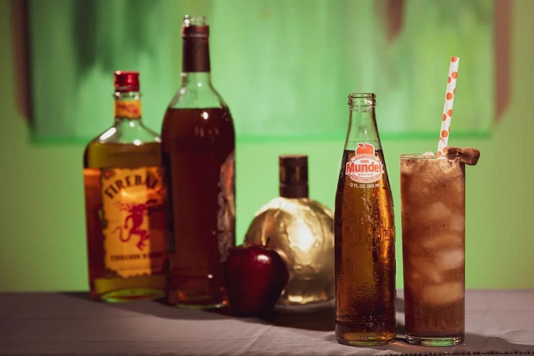 Assorted liquor bottles and an iced mixed drink with a straw arranged on a table against a green backdrop