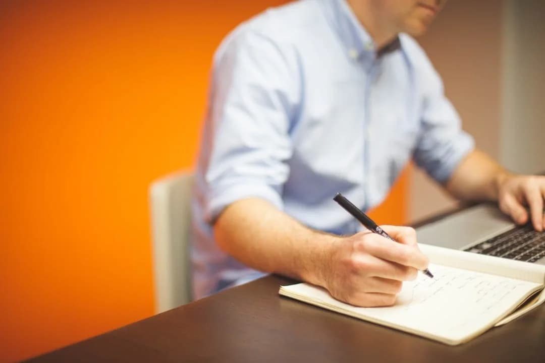 Researcher at a desk writing study notes in a notebook while working on a laptop against an orange wall