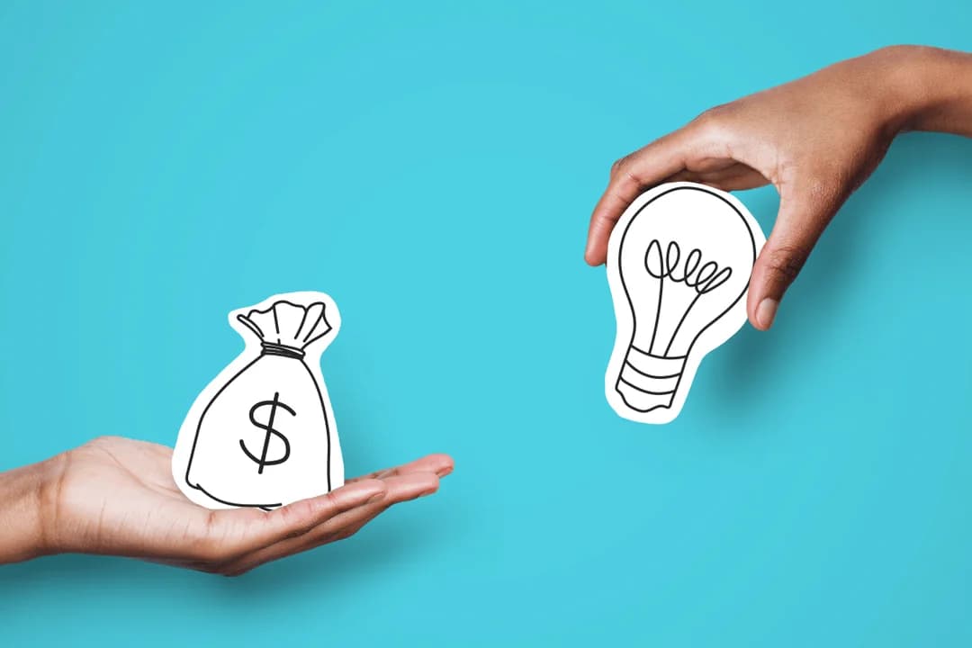 Two hands exchanging a paper money bag and a light bulb cutout on a turquoise background, symbolizing funding for ideas