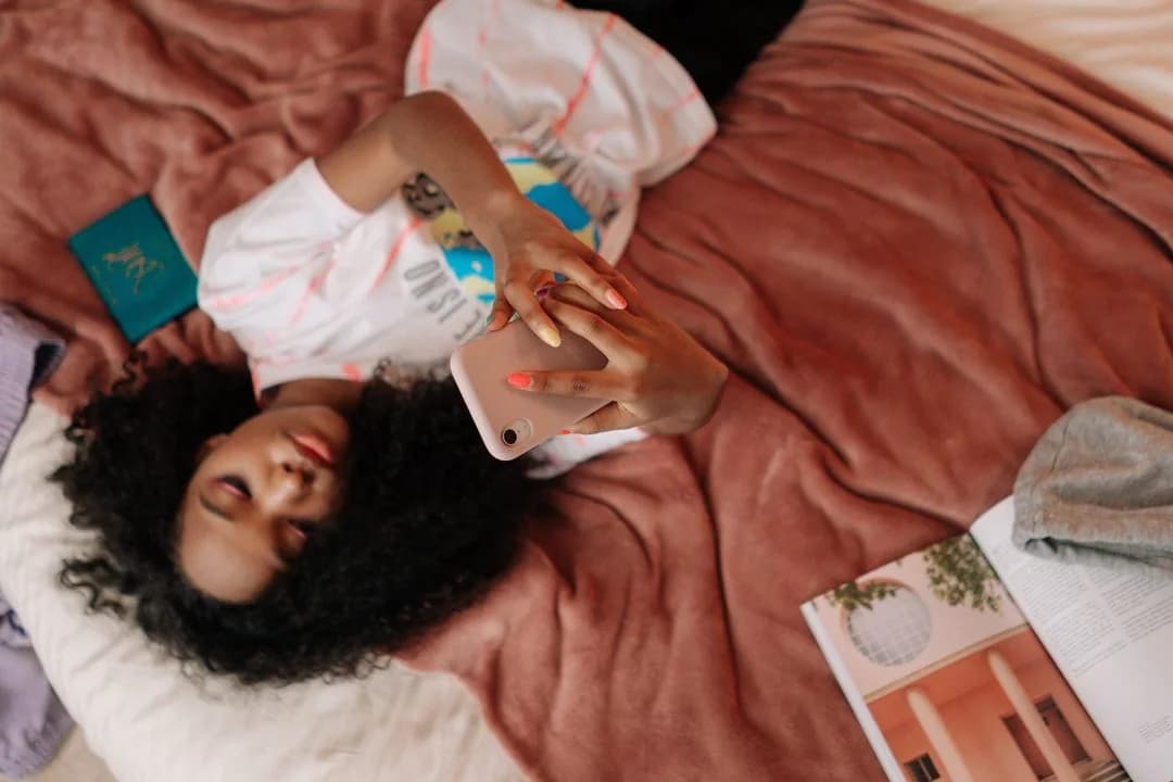 Teenage girl lying on her bed scrolling social media on her phone beside an open magazine
