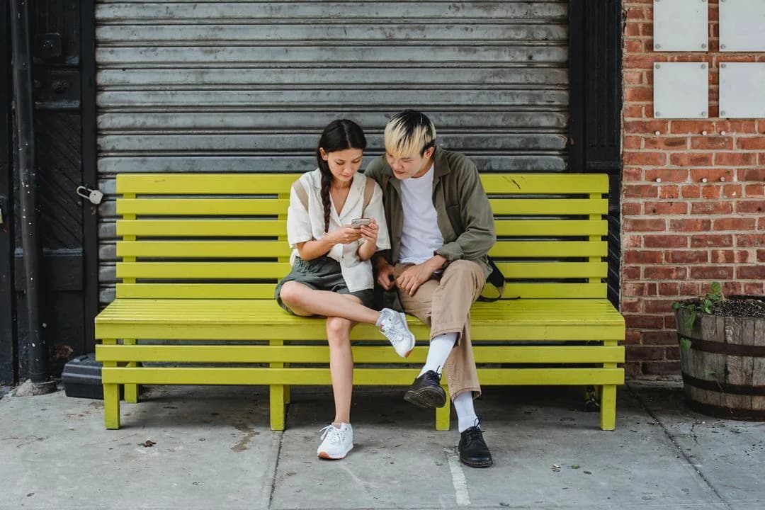 Young couple sitting close on a bright yellow street bench, looking at a smartphone together