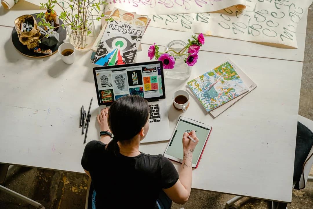 Overhead view of a researcher working across a laptop and a tablet at a desk covered in magazines and sketches