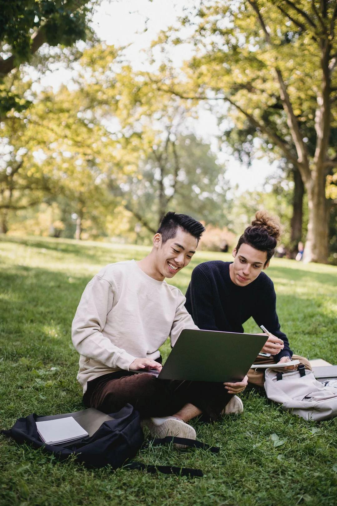 Two graduate students studying on a laptop and taking notes while sitting on the grass in a sunny park