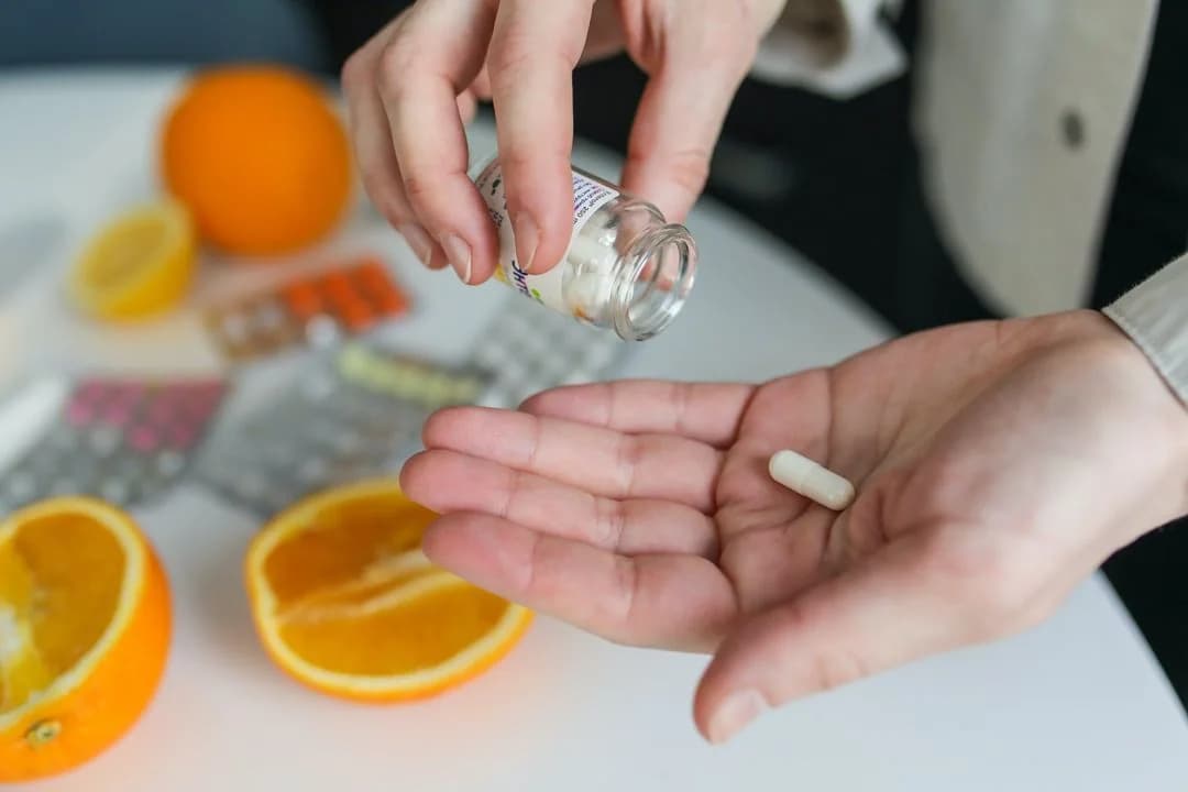 Person tipping a capsule from a medicine bottle into their palm at a table with oranges and pill packs