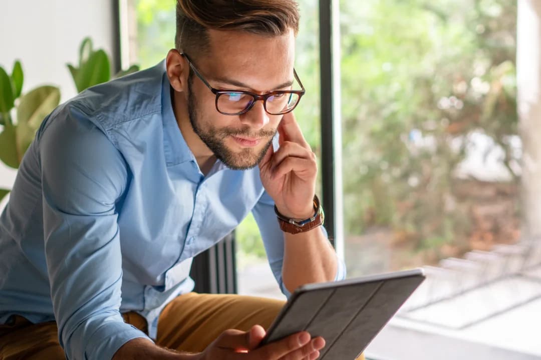 Man with glasses reviewing a survey on a tablet by a sunlit window with greenery outside