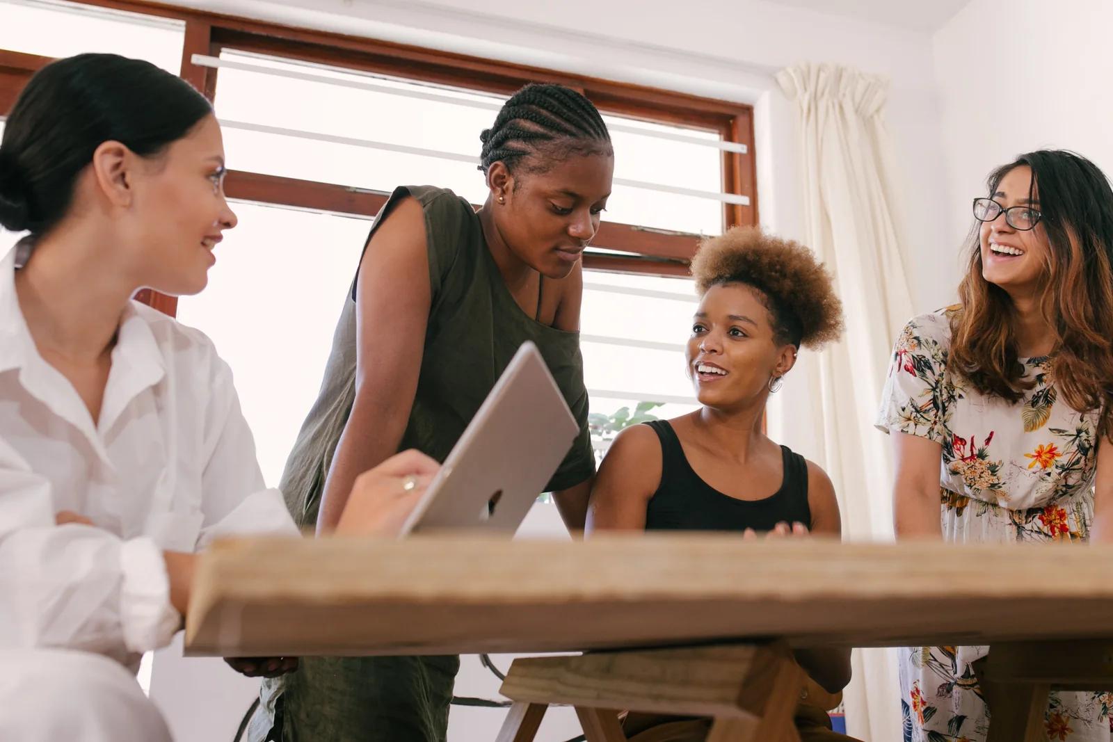 Four smiling coworkers collaborating around a laptop at a wooden table in a bright office