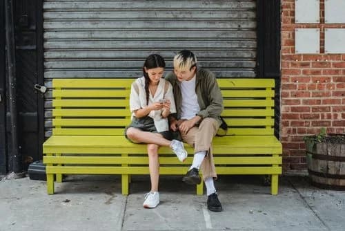Two friends on a yellow bench checking an experience sampling app on a smartphone between daily activities