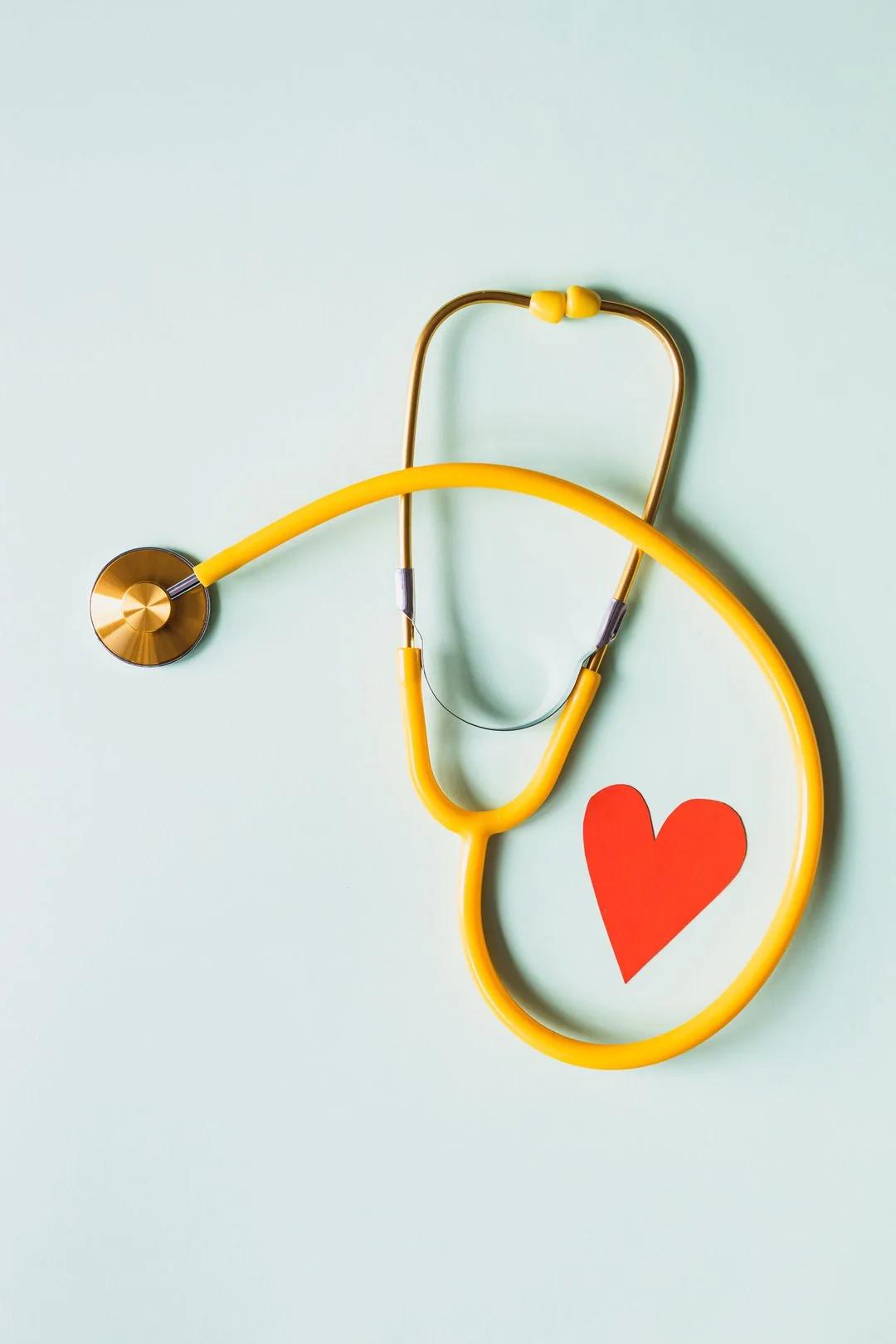 Yellow stethoscope curled around a red paper heart on a pale mint background, evoking heart health research