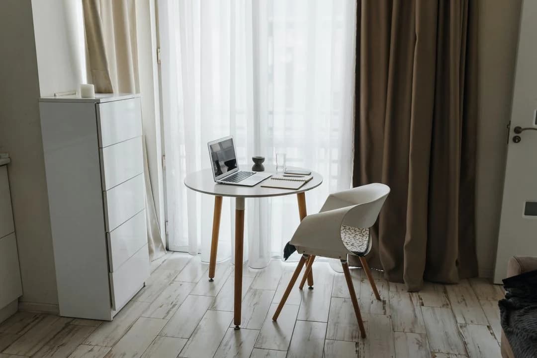Home workspace with an open laptop and notebook on a small round table beside a white chair and sheer curtains