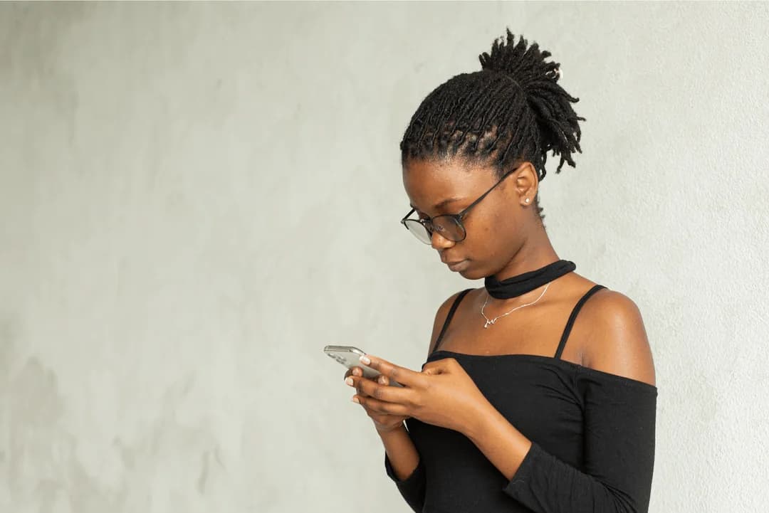 Young woman in glasses responding to a survey on her smartphone against a plain wall