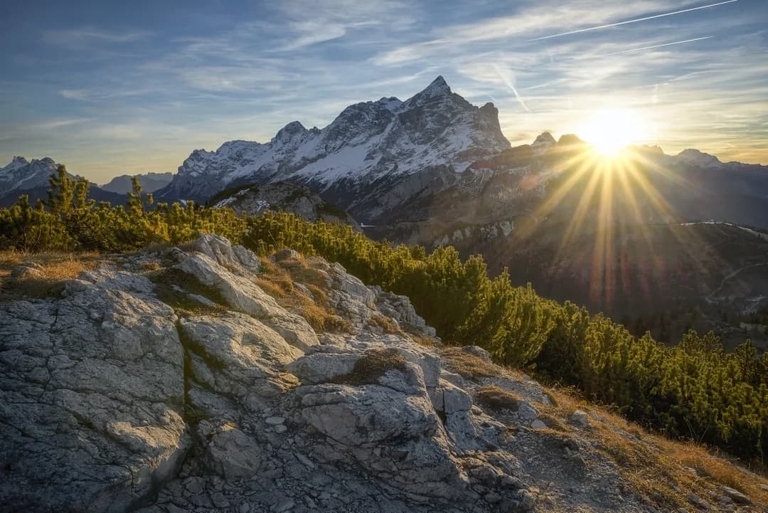 Sun rising over snow-capped mountain peaks above a rocky ridge with evergreen shrubs