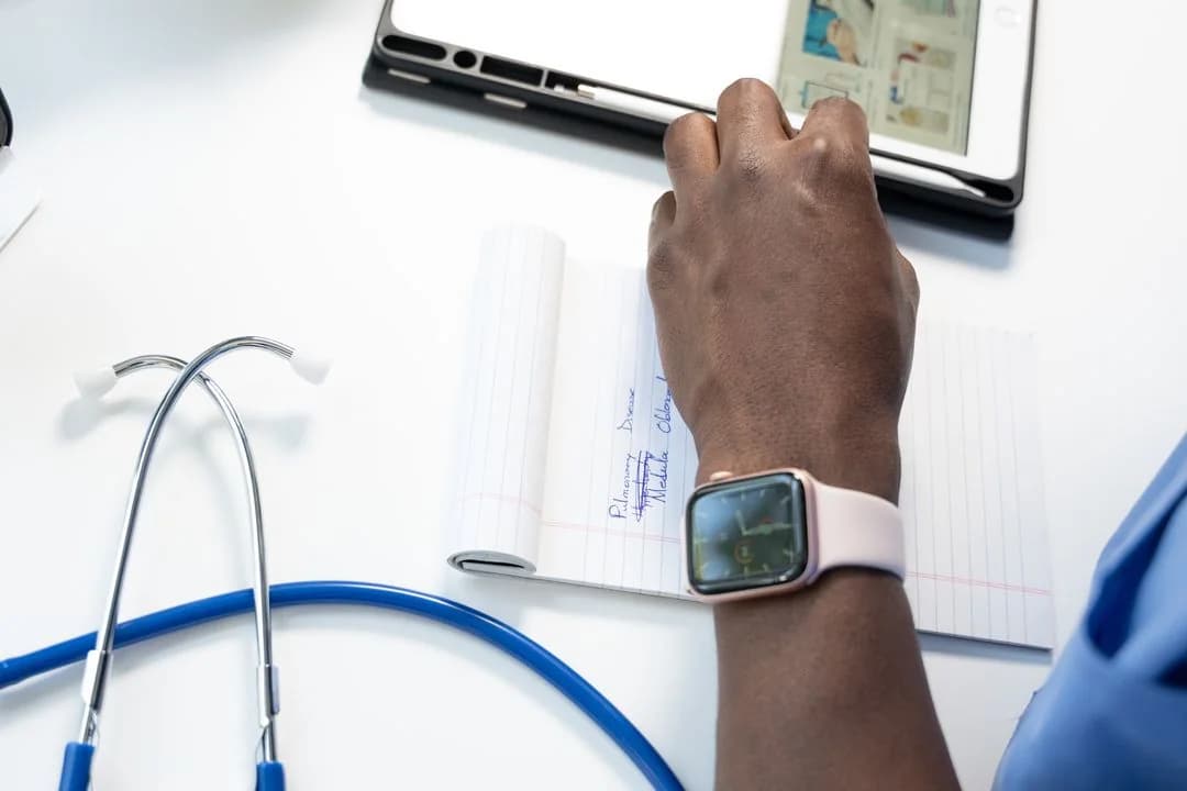 Clinician wearing a smartwatch taking notes at a desk with a stethoscope and tablet