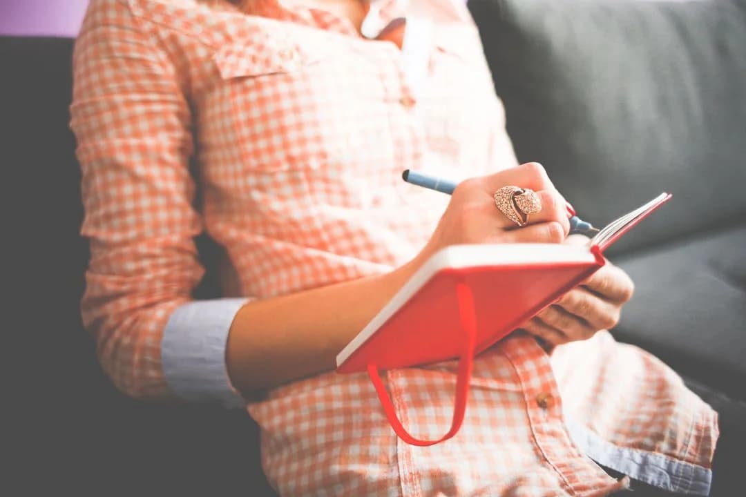 Woman on a sofa writing a diary entry with a pen in a red notebook