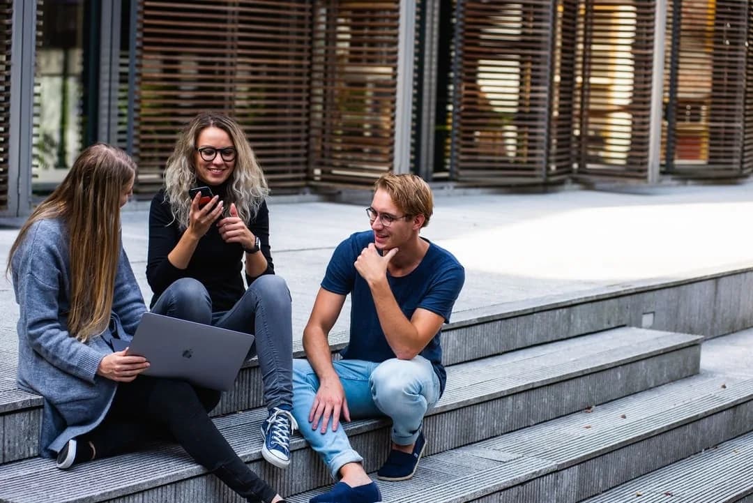 Three college students chatting on campus steps with a laptop and smartphone between classes