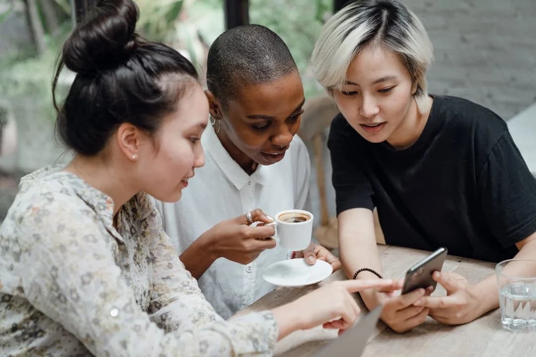 Three women at a cafe table looking together at a smartphone over coffee